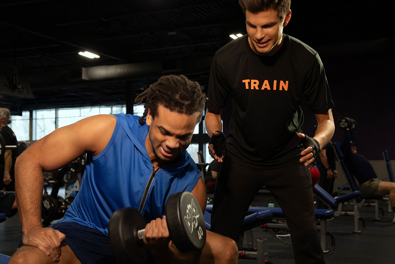 Crunch personal trainer in TRAIN shirt spotting a member during a dumbbell exercise on the gym floor