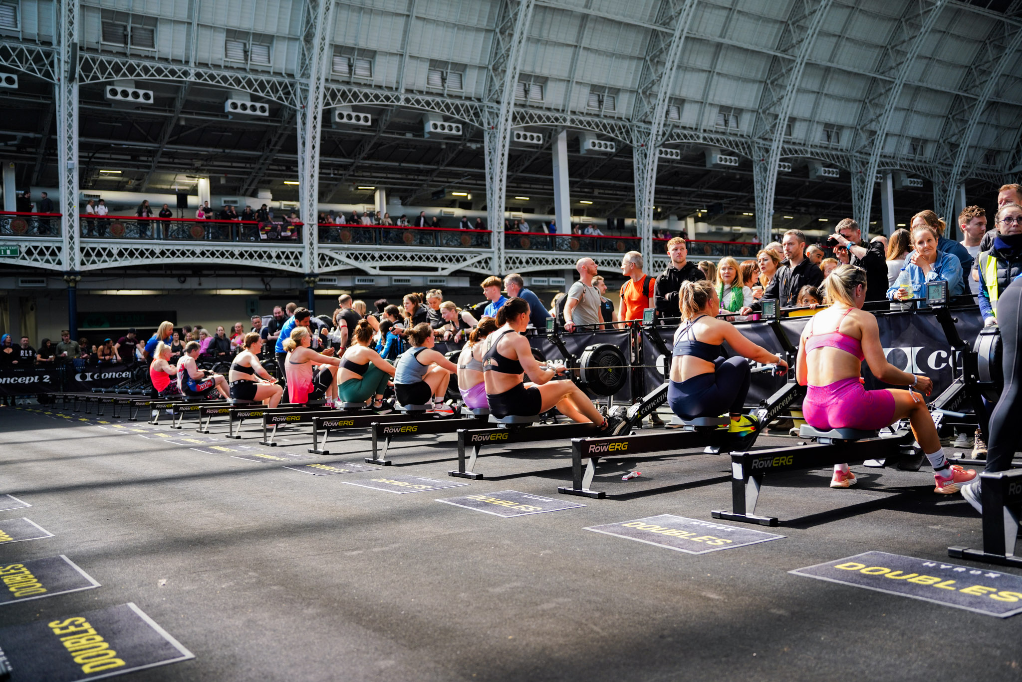 Wide view of athletes on RowERG machines at a HYROX doubles event in a Victorian exhibition hall with spectators on elevated balconies