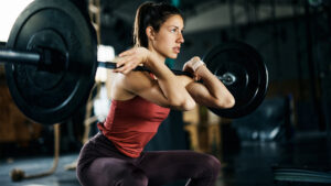 Woman performing a barbell back squat with a coach spotting and observing her form.