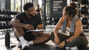 Fitness coach talking quietly with a client on the gym floor, reflecting a private conversation about medication and motivation.