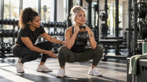 Older woman performing a loaded goblet squat with a coach spotting her in a bright strength gym